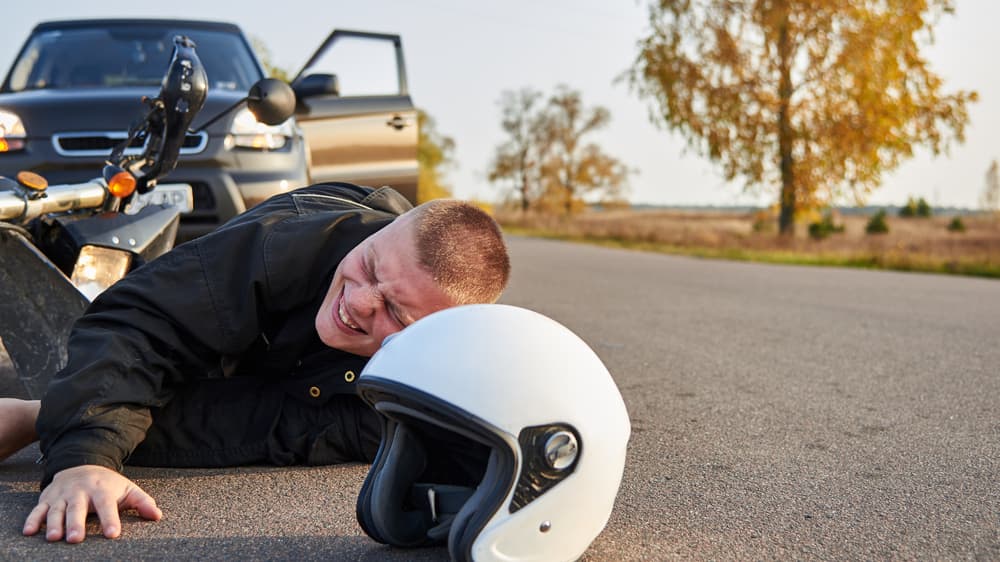 A motorcyclist lies on the asphalt beside a motorcycle and a car, suffering from severe injuries resulting from a road accident.