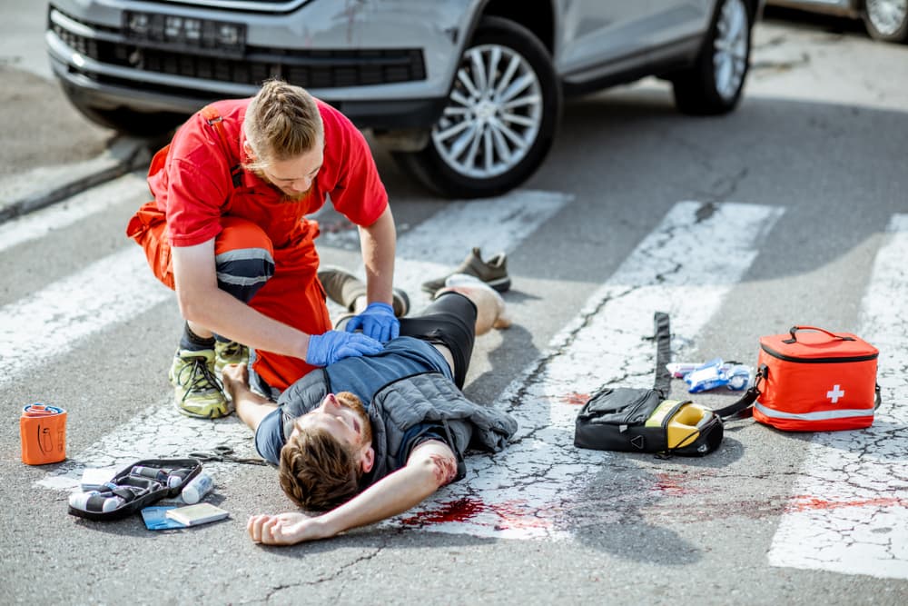 An ambulance worker is administering emergency care to an injured, bleeding man lying on the pedestrian crossing following a road accident.